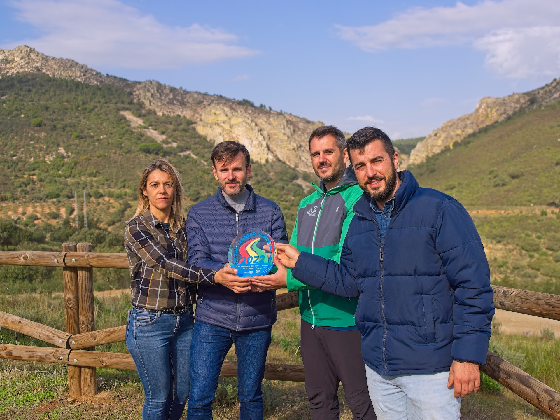 4 personas en primer plano, 1 mujer y 3 hombres, sujetan una placa conmemorativa del premio otorgado por el Mejor senderos homologado 2022. De fondo se ve la Garganta del Fraile, que es un cortado de cuarcita entre bosque mediterráneo.